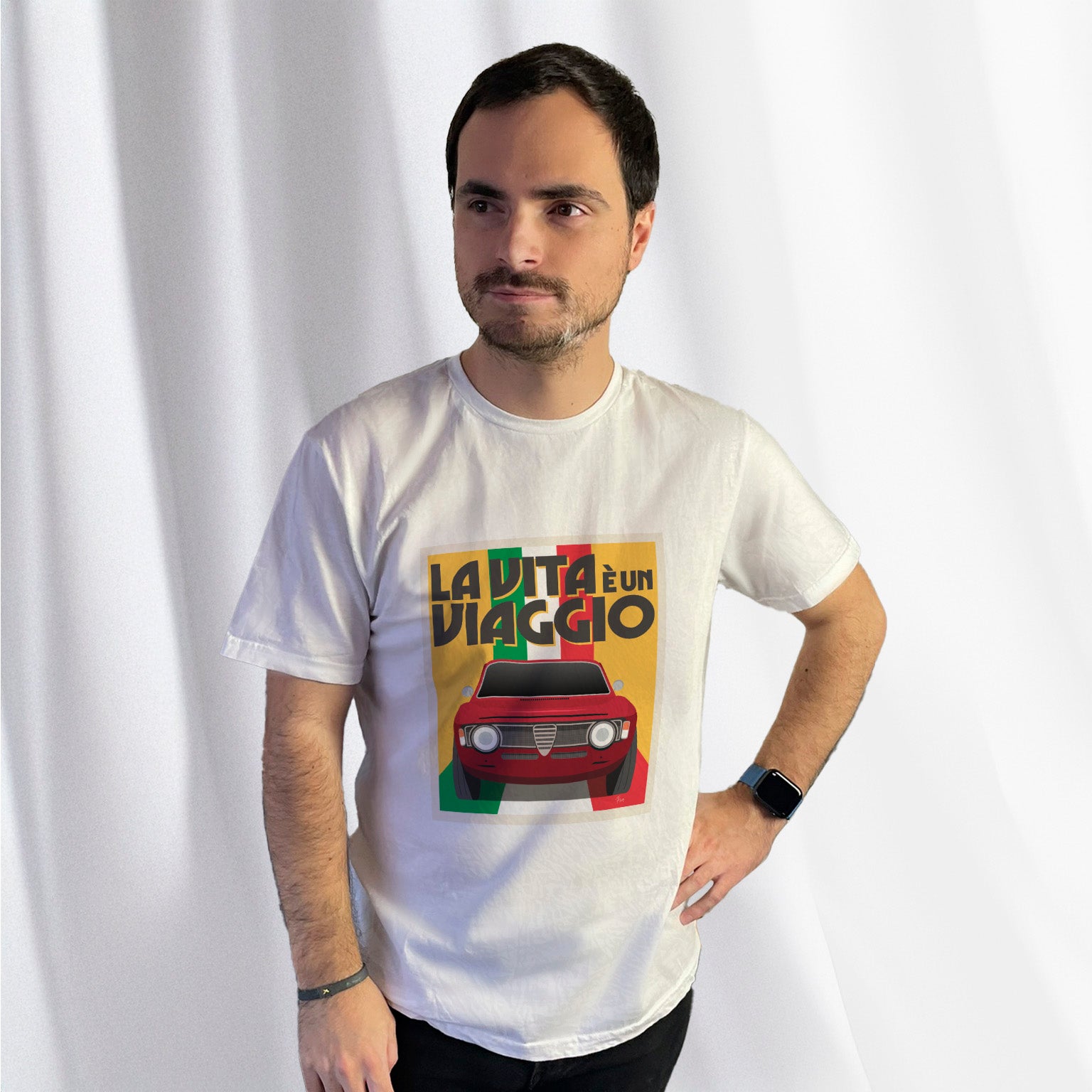 T-shirt blanc avec au milieu une illustration type affiche "La Vita à un Viaggio" reprensentant une Alfa Romeo Giulia GT sur fond jaune de face roulant sur un drapeau italien qui par du fond pour se fondre sous la voiture, porté par Corentin Gouloumy