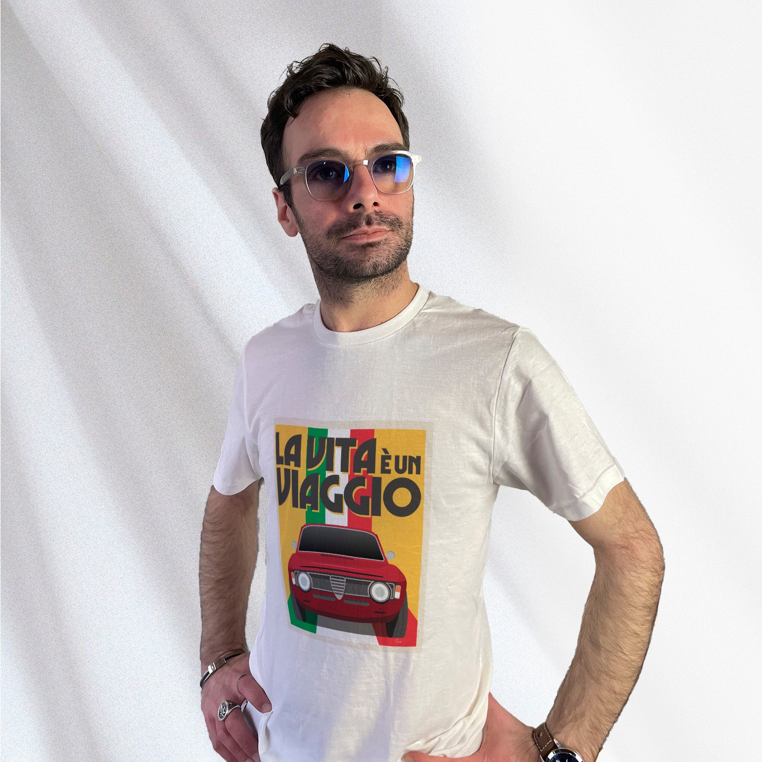 T-shirt blanc avec au milieu une illustration type affiche "La Vita à un Viaggio" reprensentant une Alfa Romeo Giulia GT sur fond jaune de face roulant sur un drapeau italien qui par du fond pour se fondre sous la voiture