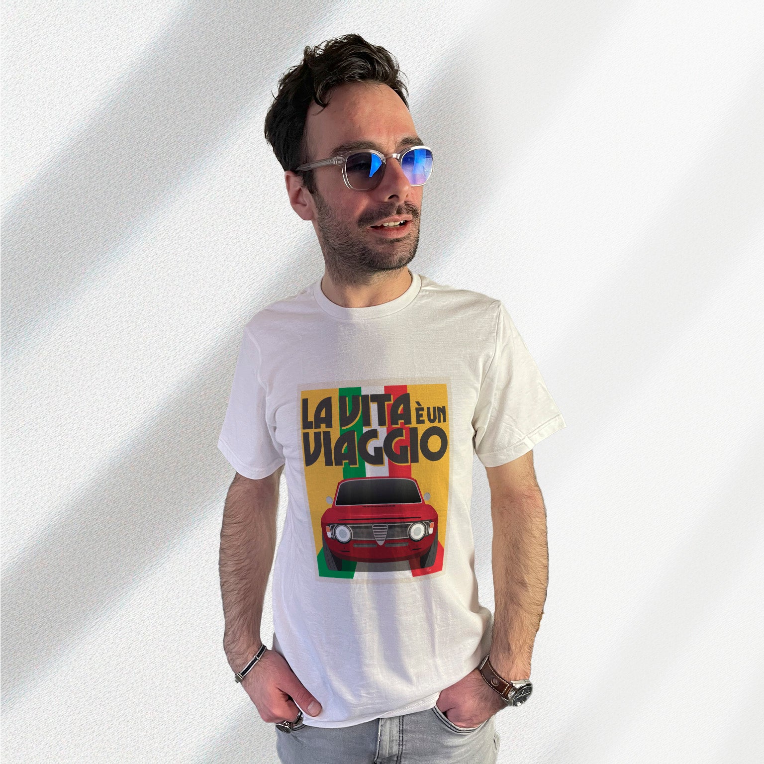 T-shirt blanc avec au milieu une illustration type affiche "La Vita à un Viaggio" reprensentant une Alfa Romeo Giulia GT sur fond jaune de face roulant sur un drapeau italien qui par du fond pour se fondre sous la voiture